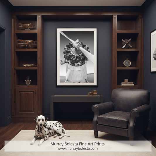 A black and white framed art print of a vintage airplane engine is displayed on a dark navy wall in a styled home office setting. A dark leather armchair sits to the right, a dalmatian lies on a light rug on the floor, and wooden shelving with decorative objects is on both sides.