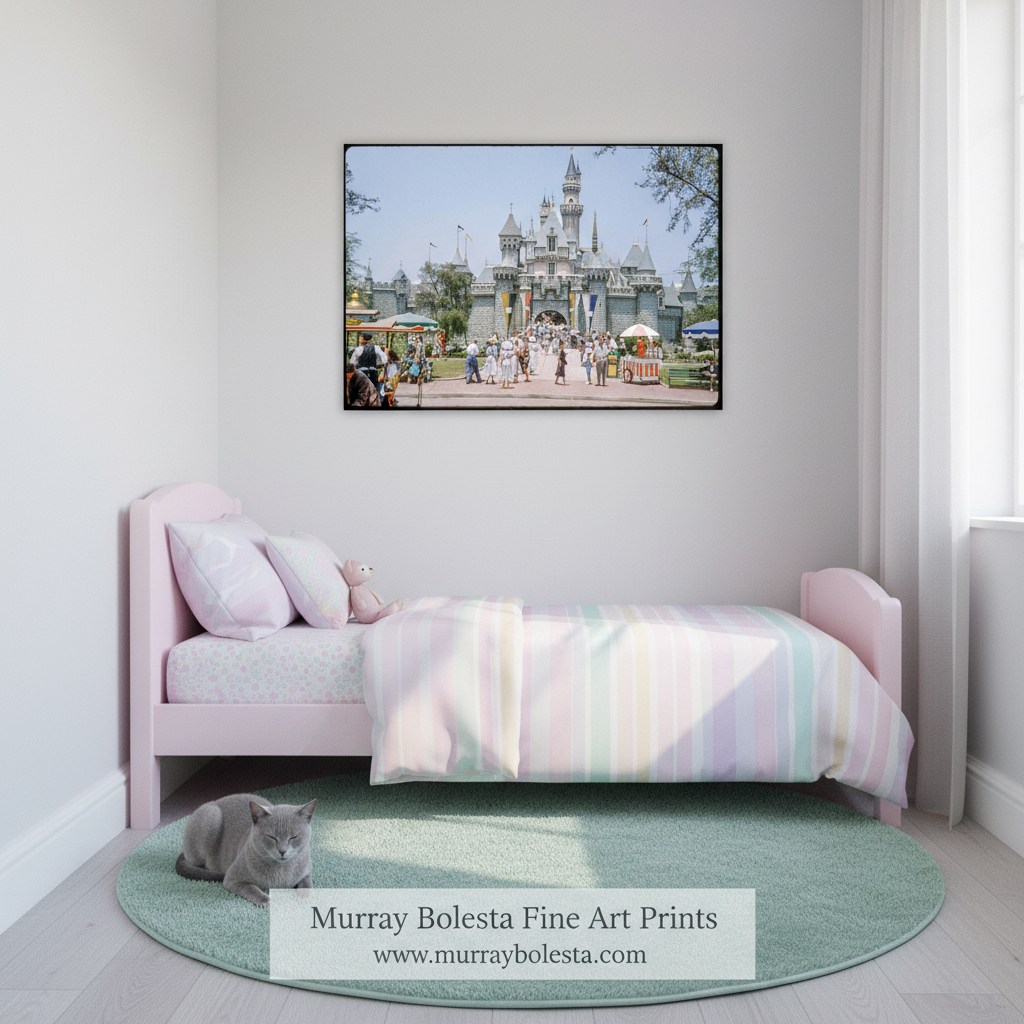 A framed art print of a fairground scene with a castle is displayed on a white wall above a light pink children's bed. A gray cat sits on a round mint green rug on the floor.