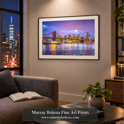 A framed art print of the Brooklyn Bridge and Manhattan skyline at dusk, displayed on a beige wall in a modern living room setting with a gray sofa and a coffee table. Art by Murray Bolesta