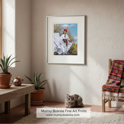 Tumacacori Fiesta. A framed art print featuring a woman in traditional white clothing with a red headpiece, hung on a textured beige wall in a styled room with potted plants, a wooden chair with a colorful blanket, and a cat on the floor. A watermark at the bottom reads 'Murray Bolesta Fine Art Prints'.