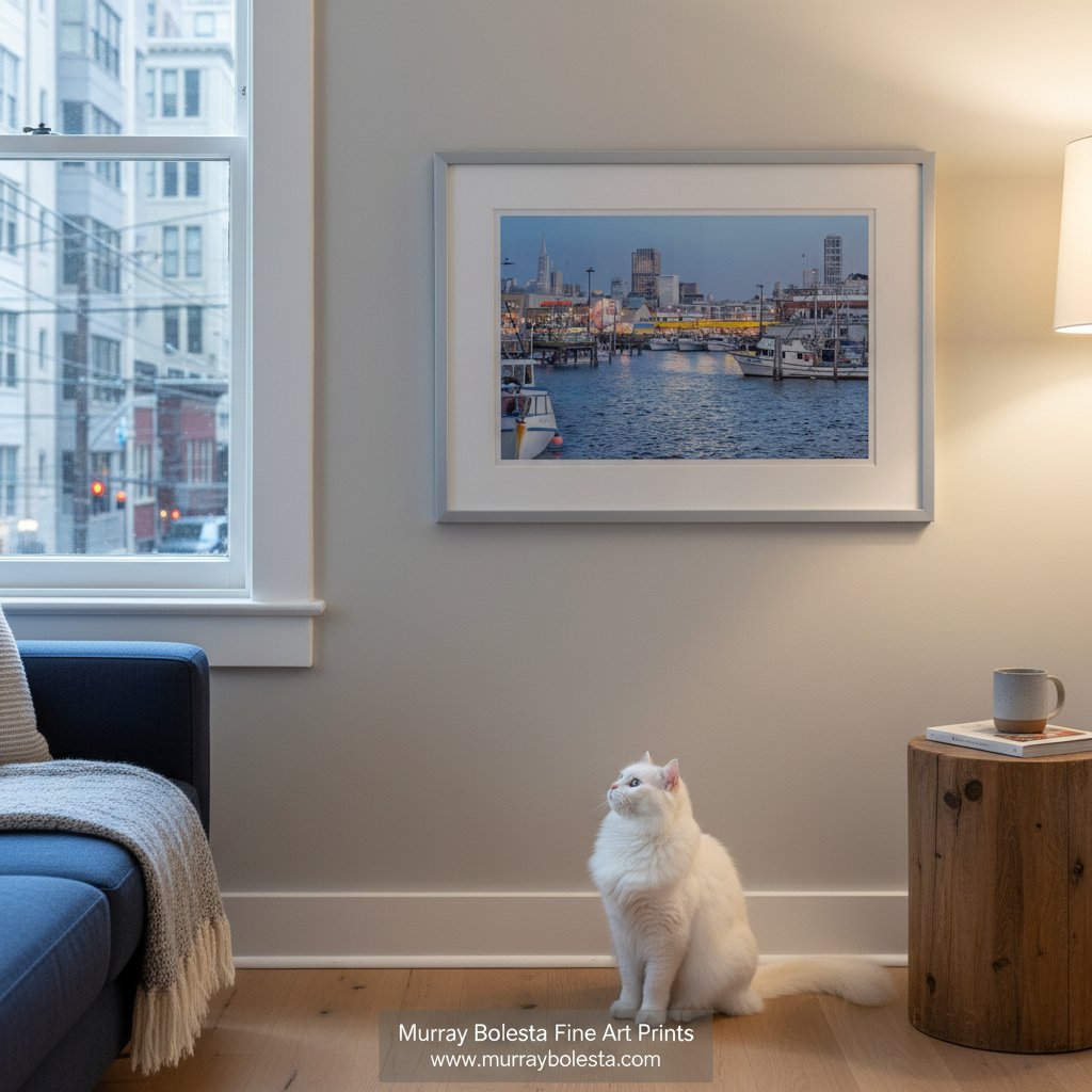 Fishermans Wharf San Francisco by Murray Bolesta! A framed art print of a harbor city scene hangs on a beige wall in a living room, with a white cat sitting below and a window and side table visible.