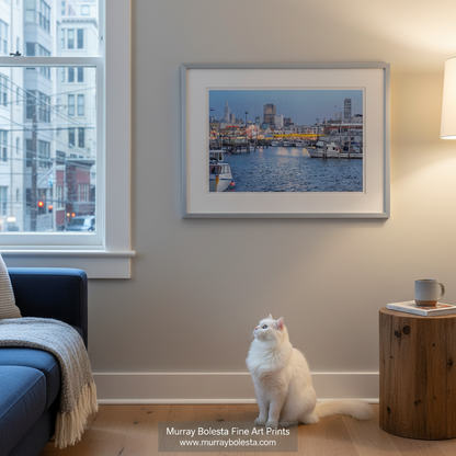 Fishermans Wharf San Francisco by Murray Bolesta! A framed art print of a harbor city scene hangs on a beige wall in a living room, with a white cat sitting below and a window and side table visible.