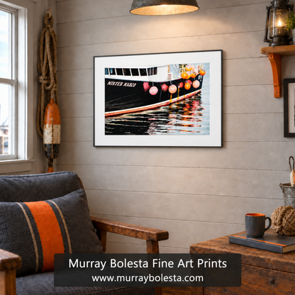 A framed art print of aboat hanging on a light gray wood-paneled wall in a cozy, rustic living room setting. The print features a black boat with orange and pink buoys and colorful reflections in the water. Boat art by Murray Bolesta