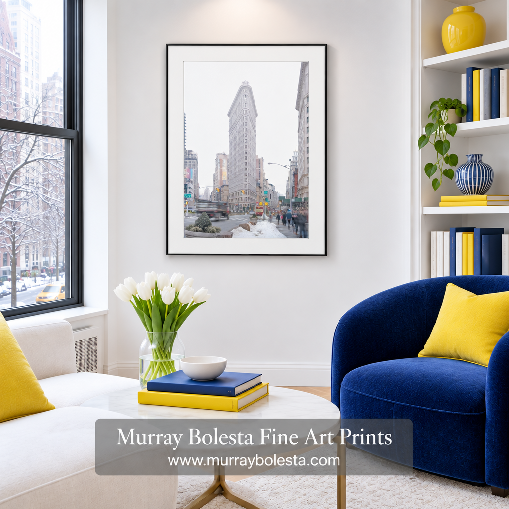 A framed art print of the Flatiron Building in New York City, shown hanging on a white wall in a modern living room setting with a blue armchair, white sofa, and yellow accents. Art by Murray Bolesta.