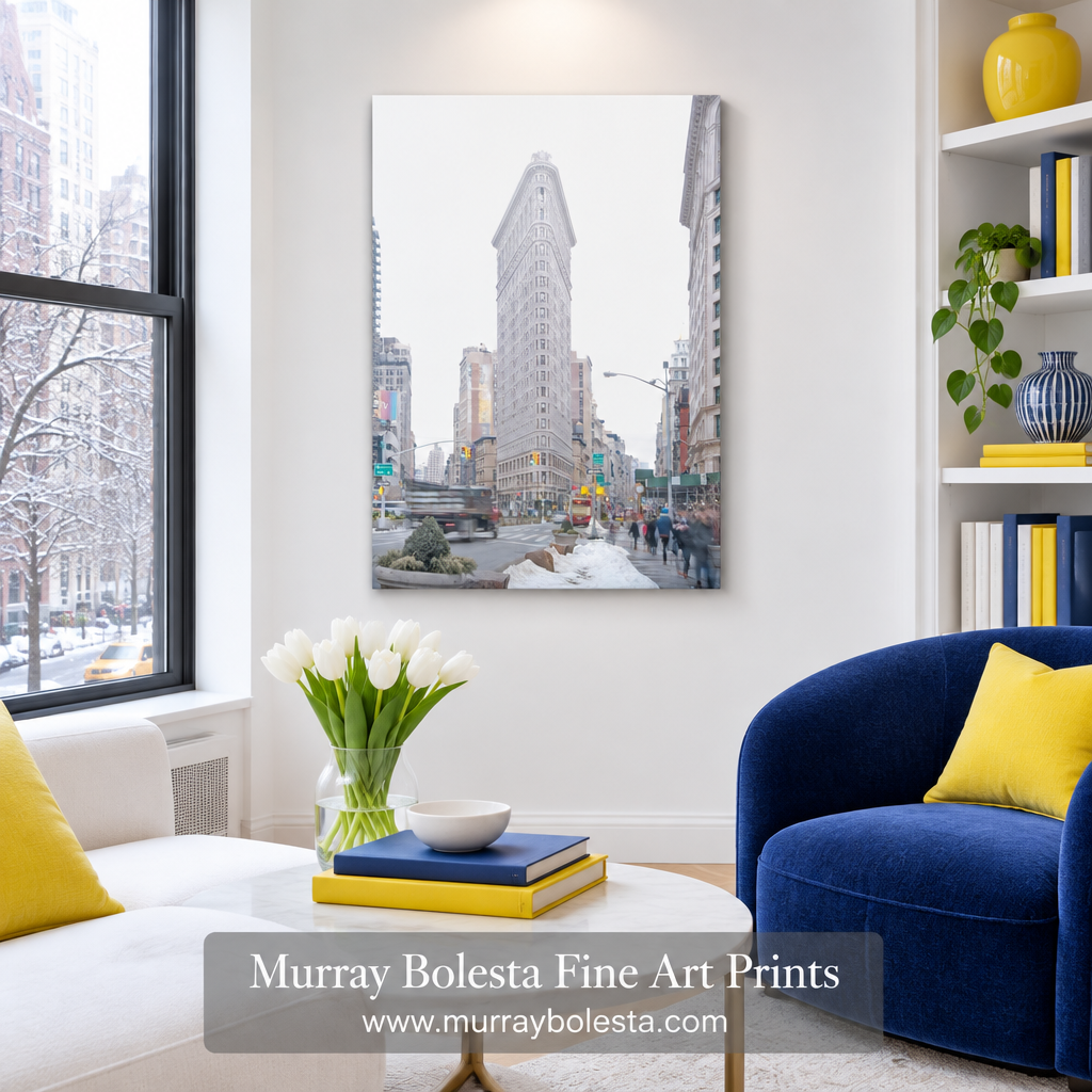 A vertical canvas art print of the Flatiron Building in New York City, shown hanging on a white wall in a modern living room setting with a blue armchair, white sofa, and yellow accents. Art by Murray Bolesta