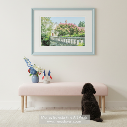 A stone bridge with a railing, surrounded by lush greenery and pink flowers, leading to a large, ornate building with a red roof and multiple towers. French Castle Photo Art Print | South of France Wall Decor by Murray Bolesta