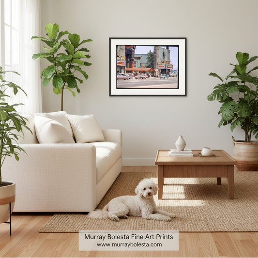 A framed art print depicting a city street scene hangs on a light beige wall in a modern, sunlit living room with a white sofa, wooden coffee table, and potted fiddle-leaf fig plants. A small white dog lies on the woven rug in front of the sofa.