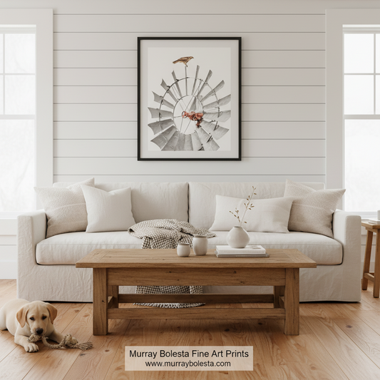 A framed wall art print featuring a stylized windmill illustration in muted gray, white, and rust tones, displayed above a white sofa in a bright living room. A small dog lies on the wood floor in the foreground, and a watermark at the bottom reads 'Murray Bolesta Fine Art Prints'.