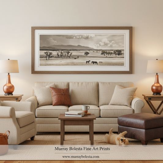 A framed sepia landscape art print by Murray Bolesta hangs on a beige wall above a light beige sofa in a styled living room. The print depicts a wide grassy plain with a few small animals under a cloudy sky.