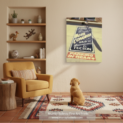 Hotel Congress Tucson by Murray Bolesta! A framed wall art print showing a vintage motel parking sign hangs on a beige wall in a styled living room. A yellow armchair, wooden shelves with decor, a patterned rug, and a small seated dog are visible.