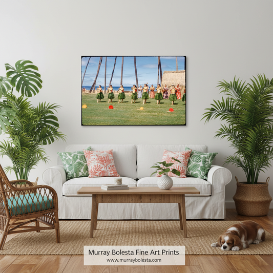 A framed art print depicting a Hawaiian hula performance on a grassy field under palm trees, displayed on a white wall above a white sofa in a styled living room with plants, a wooden coffee table, and a small dog.