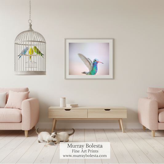 A framed art print of a hummingbird hanging on a wall in a styled living room mockup with pink armchairs, a wooden console table, and a birdcage with birds. The print itself has a white border and features a hummingbird in vibrant green, blue, and pink tones against a soft pastel background.