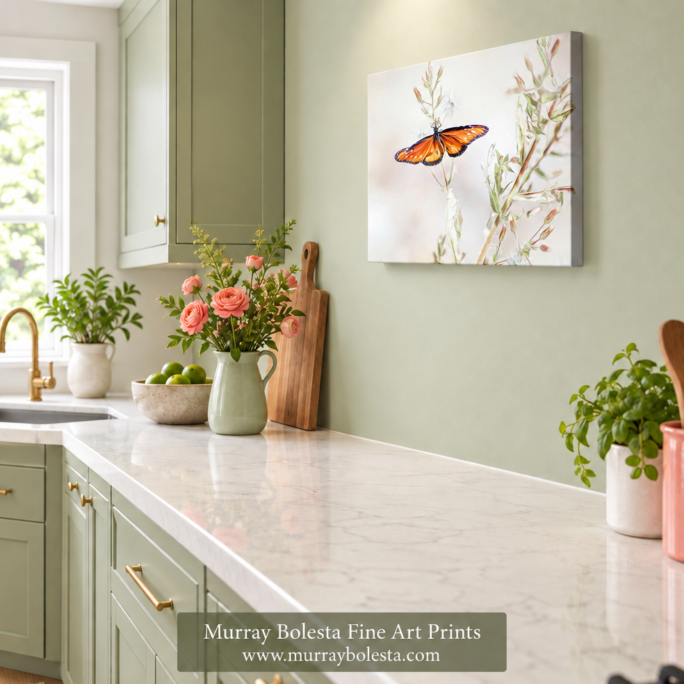 A framed wall art print showing an orange butterfly on delicate branches, displayed in a styled kitchen setting with sage green cabinets, a white marble countertop, and potted plants. A watermark at the bottom reads 'Murray Bolesta Fine Art Prints'.