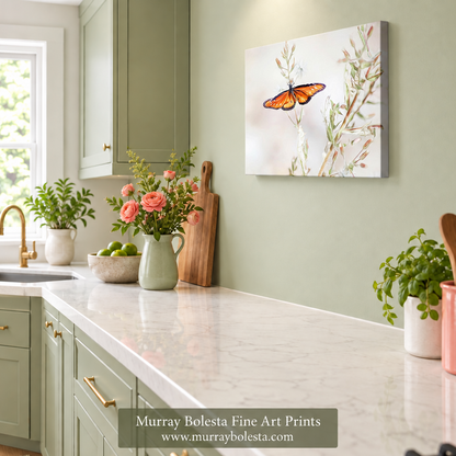 A framed wall art print showing an orange butterfly on delicate branches, displayed in a styled kitchen setting with sage green cabinets, a white marble countertop, and potted plants. A watermark at the bottom reads 'Murray Bolesta Fine Art Prints'.