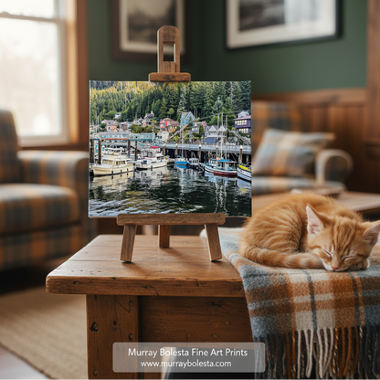 Ketchikan Alaska. Murray Bolesta. A framed art print depicting a harbor town with boats and trees, displayed on a small wooden easel on a wooden table in a cozy living room. An orange tabby cat is sleeping on a plaid throw blanket nearby.