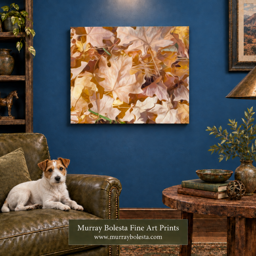 A square wall art print showing a close-up of overlapping autumn leaves in cream, tan, brown, and muted gold tones, displayed above a brown leather sofa in a styled living room. A white and tan dog lies on the couch, and a side table with books and a lamp is visible to the right. By Murray Bolesta.