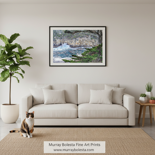 Point Lobos California! A framed landscape art print by Murray Bolesta hangs on a beige wall above a light beige sofa in a styled living room. The print depicts a rocky coastal cliff with crashing waves and green trees.