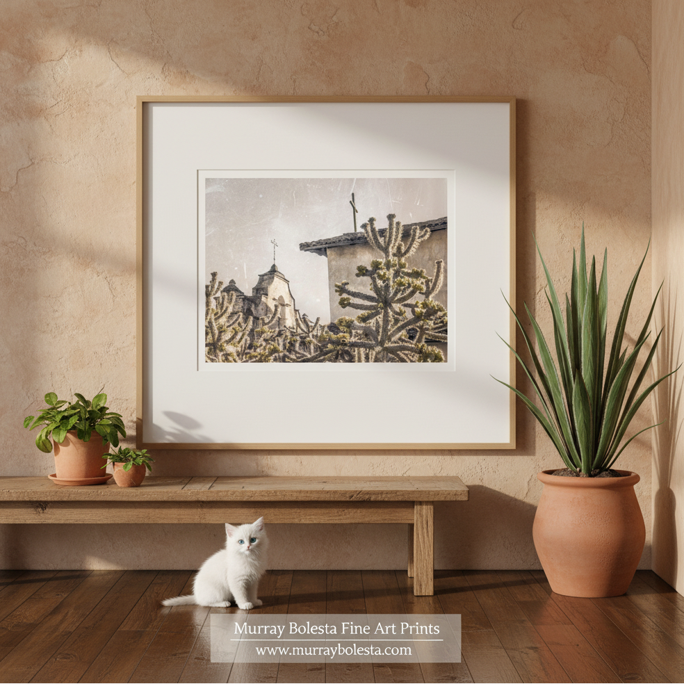 Lompoc California Spanish Mission by Murray Bolesta! A framed art print featuring a sepia-toned desert scene with cacti and a building, displayed on a beige wall above a wooden bench. A white kitten sits on the floor below, with potted plants on both sides.