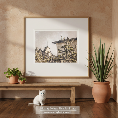 Lompoc California Spanish Mission by Murray Bolesta! A framed art print featuring a sepia-toned desert scene with cacti and a building, displayed on a beige wall above a wooden bench. A white kitten sits on the floor below, with potted plants on both sides.