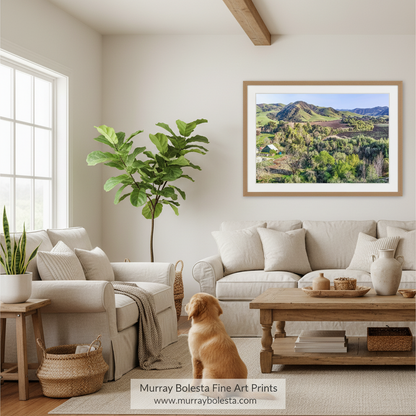 Lompoc California farm land! A framed landscape art print of green hills and mountains under a blue sky, displayed on a beige wall in a bright, modern living room with a dog sitting on the floor. The print is branded with 'Murray Bolesta Fine Art Prints' at the bottom.