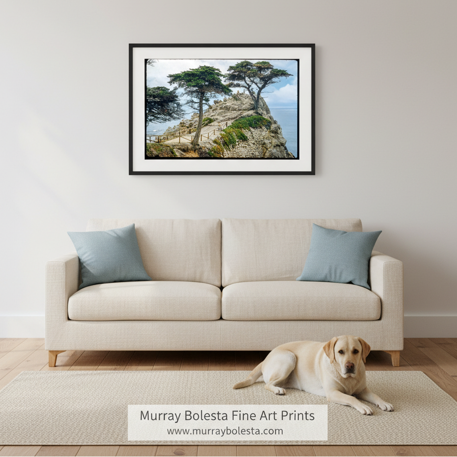 A framed landscape art print of coastal cypress trees on a rocky cliffside with the ocean, displayed on a light gray wall above a cream sofa with blue pillows. A yellow Labrador retriever is lying on a beige rug on the floor in front of the sofa.