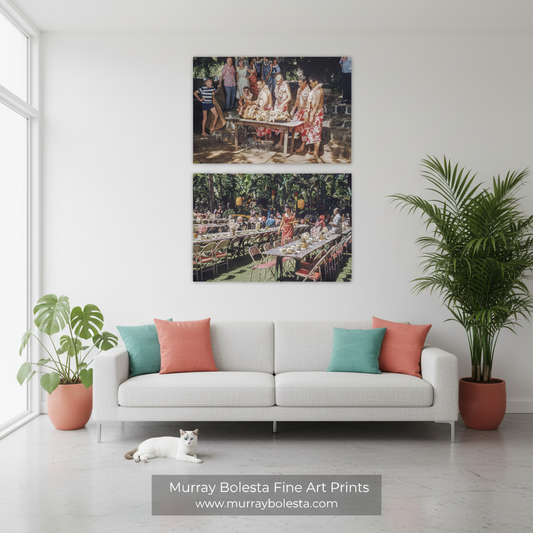 A styled living room with a white sofa, teal and coral pillows, a white cat on the floor, and two framed art prints hanging above the couch. The prints depict outdoor dining scenes.