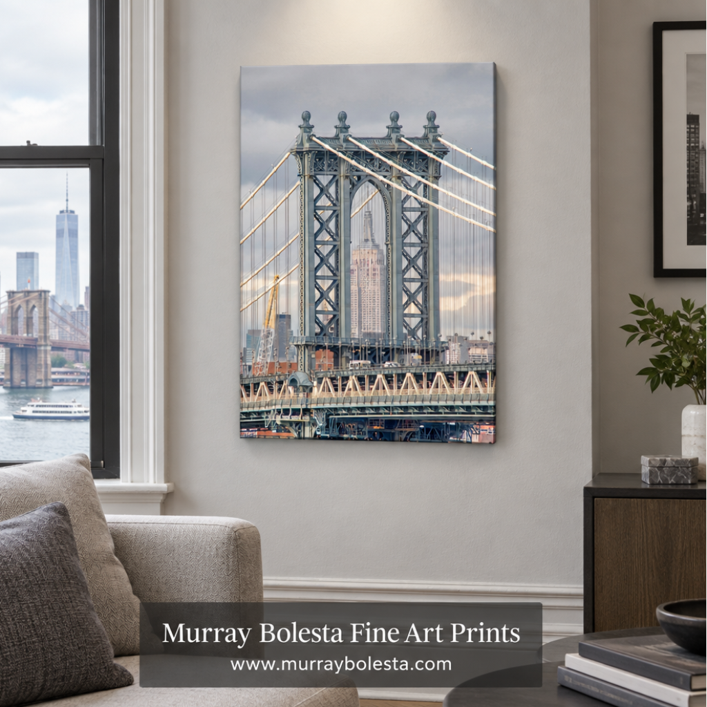 A vertical canvas wall art print depicting the Manhattan Bridge over the water with the New York City skyline, displayed on a light gray wall in a modern living room setting. A watermark banner for 'Murray Bolesta Fine Art Prints' is visible at the bottom.