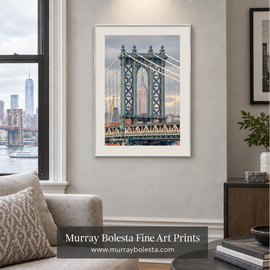 A framed art print of the Manhattan Bridge in New York City, displayed on a light gray wall in a modern living room setting. The print features a white frame and is shown above a beige sofa with a window view of the Brooklyn Bridge and skyline. Art by Murray Bolesta.