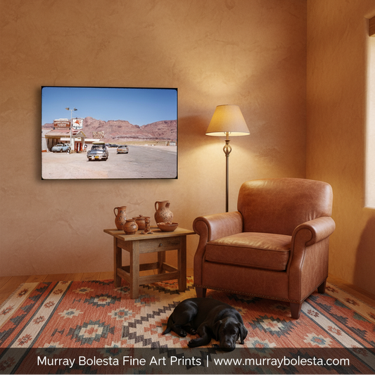 Marble Canyon, Murray Bolesta. A framed art print depicting a desert gas station scene is displayed on a textured tan wall in a styled living room mockup with a brown leather armchair, a wooden side table, a lamp, and a black dog on a patterned rug. Text at the bottom reads 'Murray Bolesta Fine Art Prints | www.