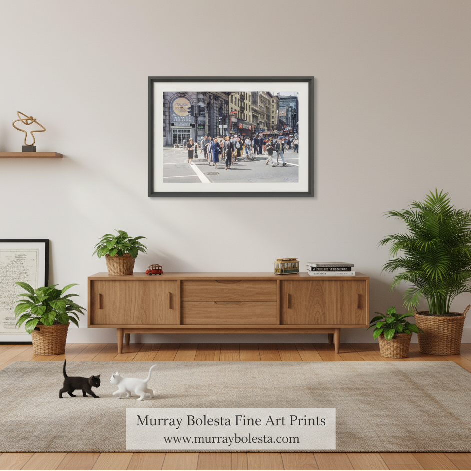 A framed art print depicting a busy city street scene hangs on a light beige wall above a mid-century modern wooden credenza in a styled living room. The print is from Murray Bolesta Fine Art Prints, as indicated by the text overlay at the bottom.