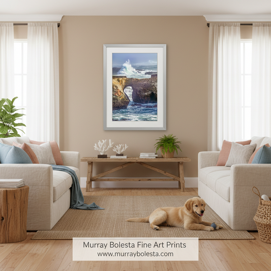 A framed art print of a coastal rock formation with crashing waves, displayed on a beige wall in a modern living room setting with two cream sofas, a wooden coffee table, and a golden retriever lying on the floor. A banner at the bottom reads 'Murray Bolesta Fine Art Prints'.
