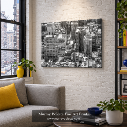A black and white aerial photograph of a dense city skyline, displayed as a wall art print in a modern living room setting with a gray sofa, yellow pillow, and brick wall. Midtown NYC winter art by Murray Bolesta