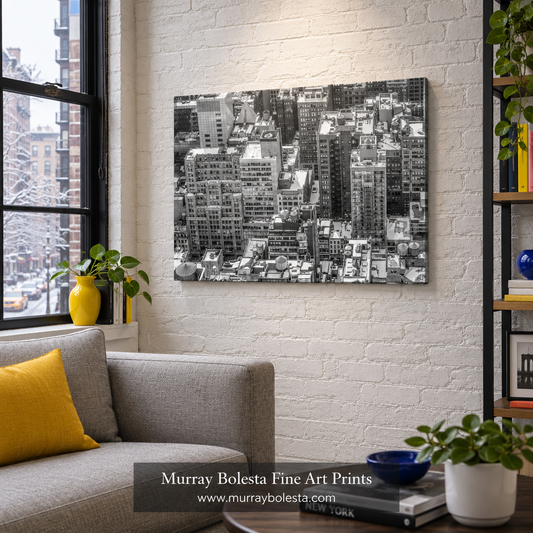A black and white aerial photograph of a dense city skyline, displayed as a wall art print in a modern living room setting with a gray sofa, yellow pillow, and brick wall. Midtown NYC winter art by Murray Bolesta