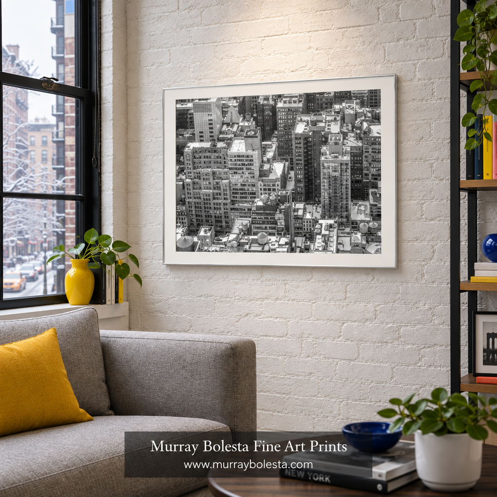 A framed black and white cityscape art print hangs on a white brick wall in a modern living room setting with a gray sofa, yellow accent pillow, and potted plants. NYC winter art by Murray Bolesta.