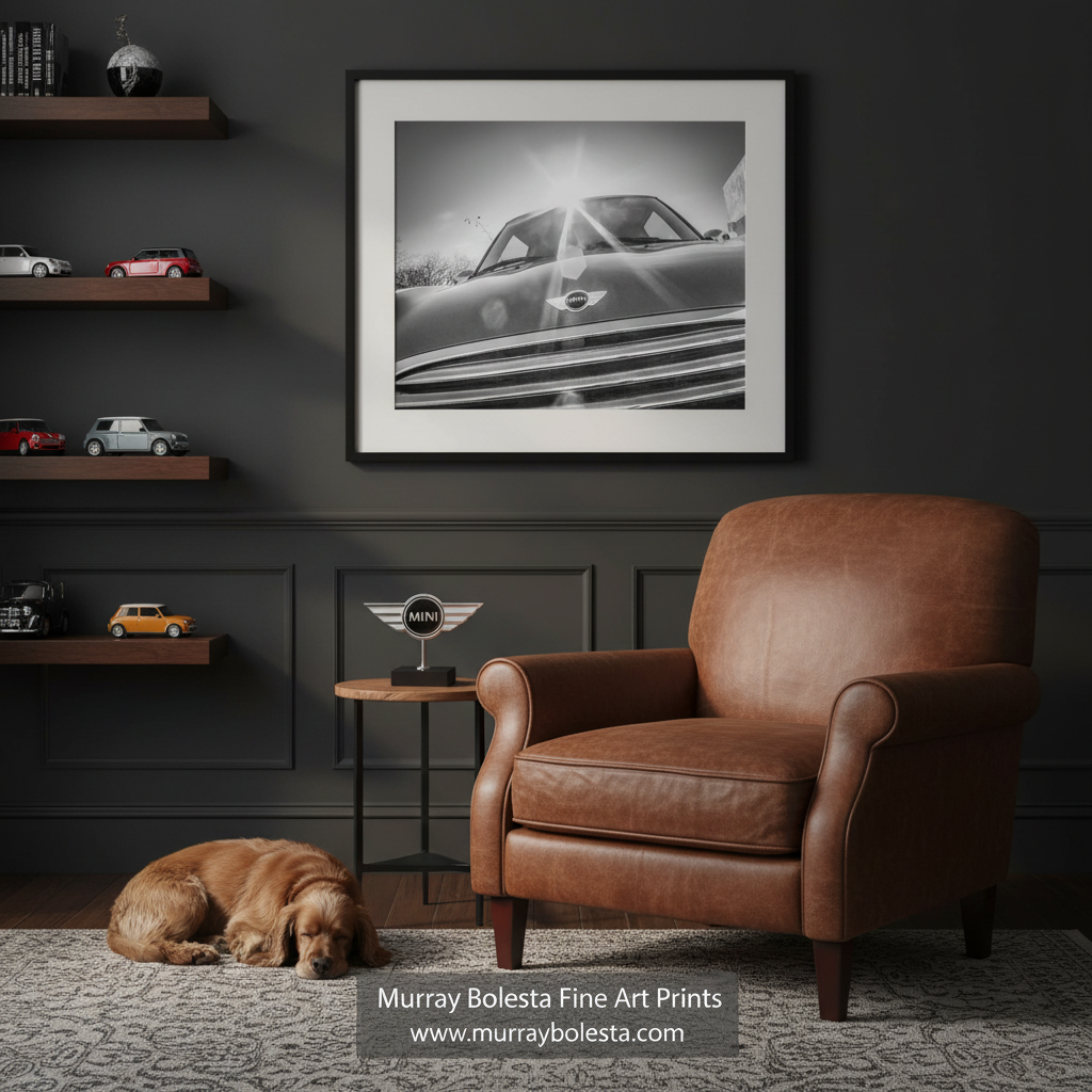 Mini Cooper car by Murray Bolesta. A framed black and white art print of a classic car is displayed on a dark gray wall in a styled room with a brown leather armchair, a dog on the floor, and floating shelves with toy cars. A watermark at the bottom reads 'Murray Bolesta Fine Art Prints'.