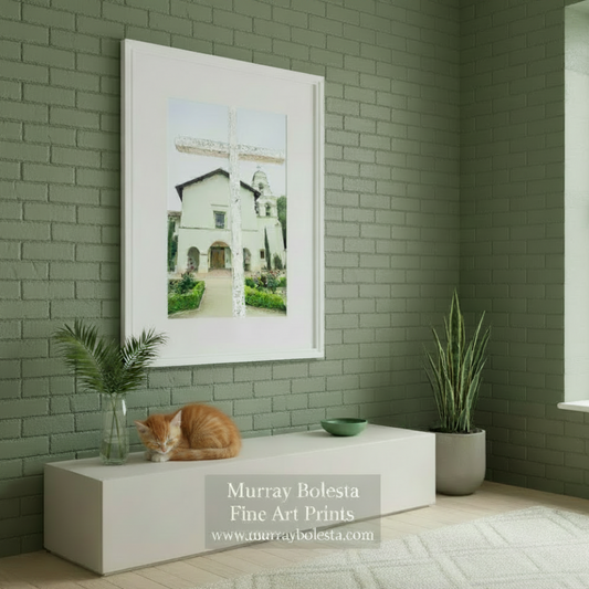 A white cross stands in front of a white building with a bell tower, surrounded by a well-maintained garden with colorful flowers. Mission San Juan Bautista Art Print – California Historic Church Photo by Murray Bolesta