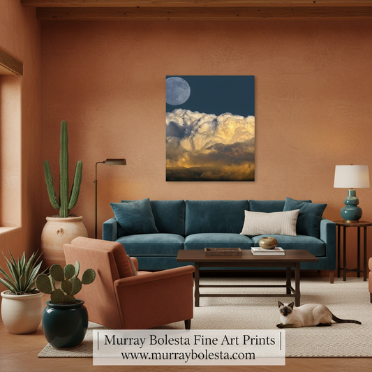 A framed wall art print showing a night sky with a large moon and dramatic clouds in blue, white, and gold tones, displayed above a teal sofa in a styled living room. A cat lies on the rug in front, and a watermark at the bottom reads 'Murray Bolesta Fine Art Prints www.