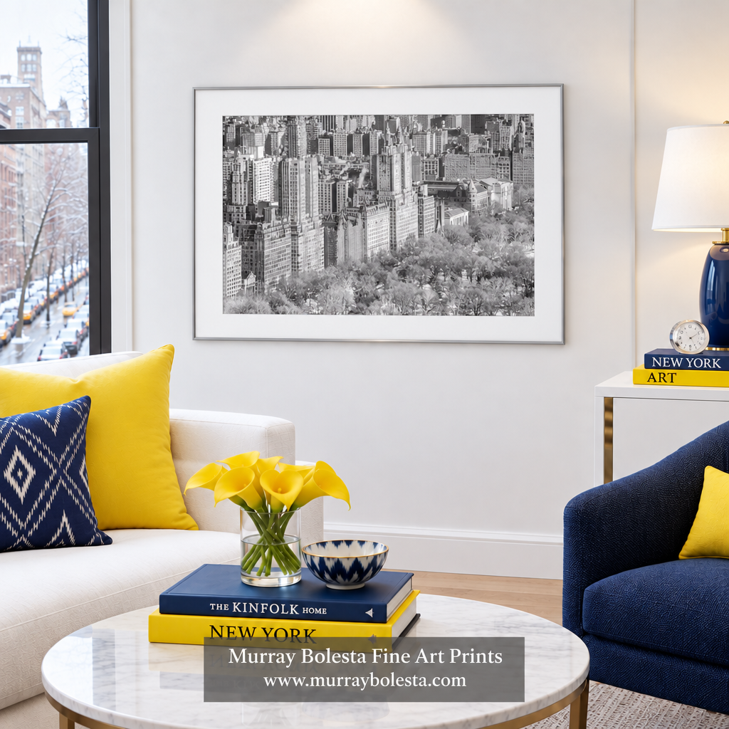 A framed black and white art print of a city skyline hangs on a white wall in a modern living room setting with yellow accent pillows, a marble coffee table, and decorative books. Upper west side NYC art by Murray Bolesta