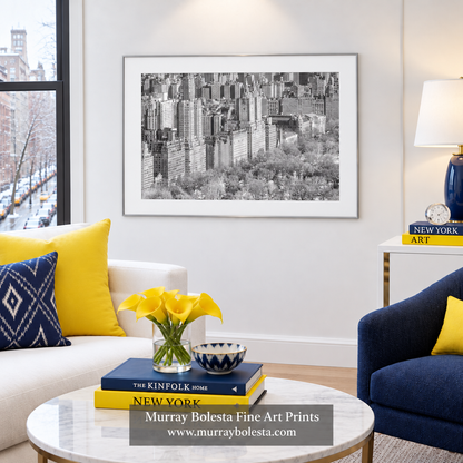 A framed black and white art print of a city skyline hangs on a white wall in a modern living room setting with yellow accent pillows, a marble coffee table, and decorative books. Upper west side NYC art by Murray Bolesta