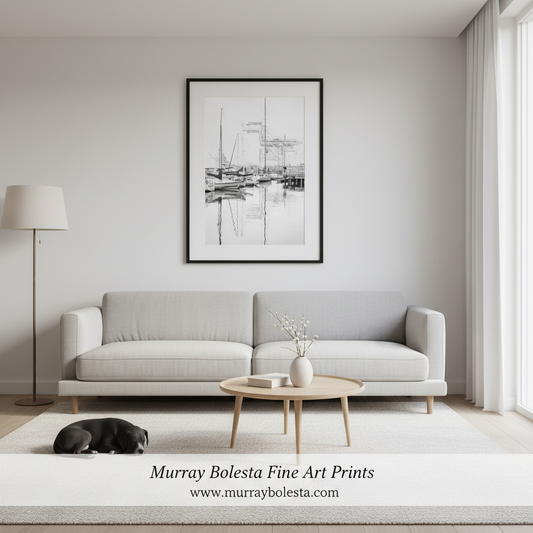 A black and white framed art print depicting a harbor scene, displayed on a light gray wall above a modern light gray sofa in a minimalist living room. A black cat sleeps on a white rug on the floor, and a wooden coffee table sits in front.