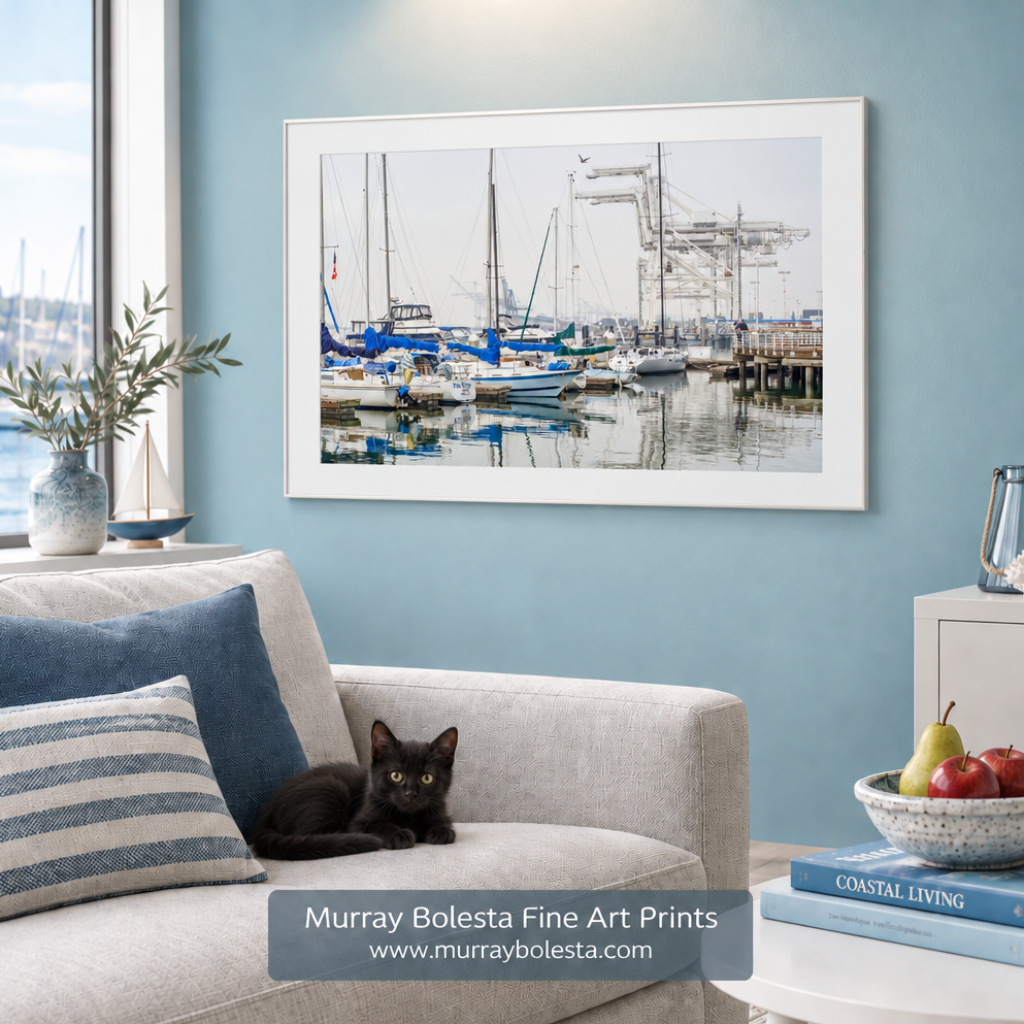 A framed art print of a harbor scene with sailboats and cranes hangs on a light blue wall above a beige sofa with blue and striped pillows. A black cat rests on the sofa, and a side table nearby holds fruit and books. Oakland Marina boats art by Murray Bolesta