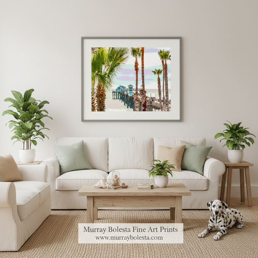 Oceanside Ca. A framed art print depicting a tropical beach scene with palm trees and a pier, displayed on a light beige wall above a white sofa in a styled living room. The print is by Murray Bolesta, whose brand name and website are displayed at the bottom of the image.