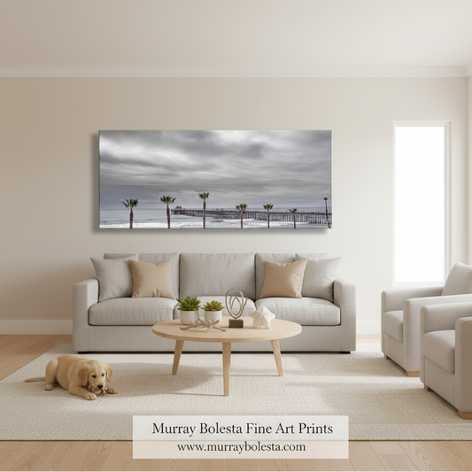Oceanside CA. A large horizontal canvas print of a pier over the ocean under a cloudy sky, displayed on a beige wall above a light gray sofa in a modern living room. A small dog is lying on the rug in the foreground, and a watermark at the bottom reads 'Murray Bolesta Fine Art Prints'.