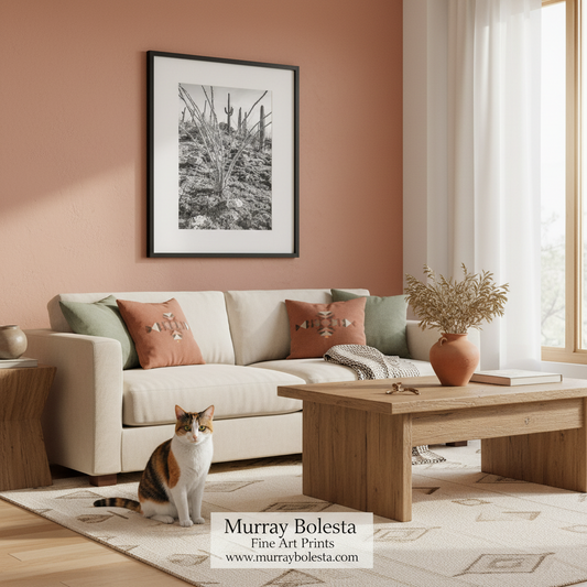 A framed black and white art print by Murray Bolesta hangs on a terracotta-colored wall above a cream sofa in a styled living room with a cat on the rug and a wooden coffee table.