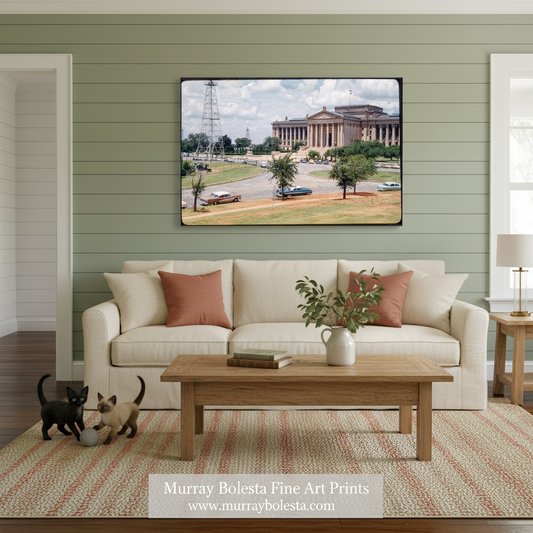 A framed art print depicting a classical American building scene is displayed on a sage green paneled wall above a cream sofa in a styled living room. The print is the product being sold, and the room serves as a lifestyle mockup context.