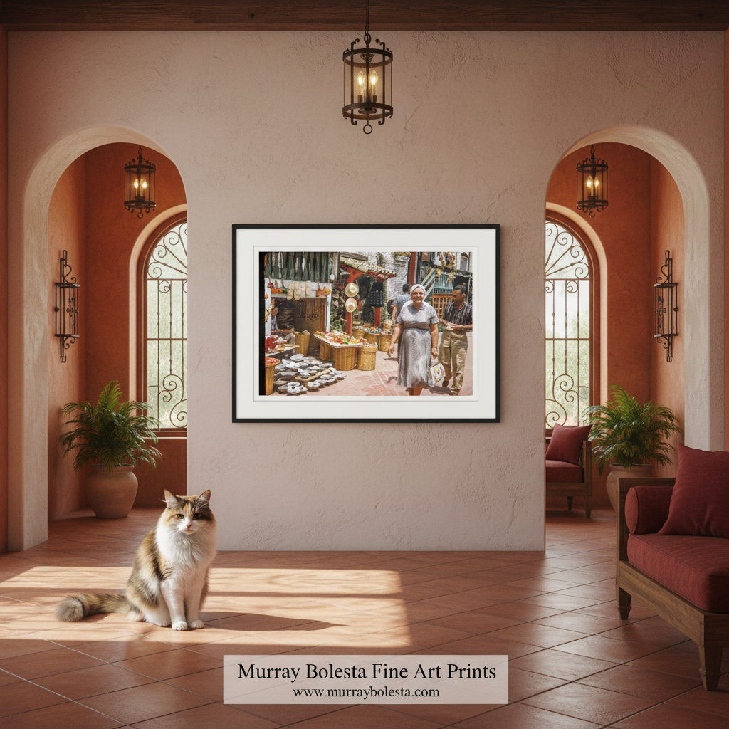 A framed art print depicting a bustling market scene with a woman in a white dress, displayed on a beige wall in a warm, terracotta-colored room with arches, a cat on the floor, and a watermark at the bottom reading 'Murray Boleta Fine Art Prints'.