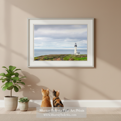 A white lighthouse stands on a grassy hill overlooking the ocean, with a cloudy sky in the background. Yaquina Head Lighthouse Art | Oregon Coast Minimalist Ocean Print by Murray Bolesta!