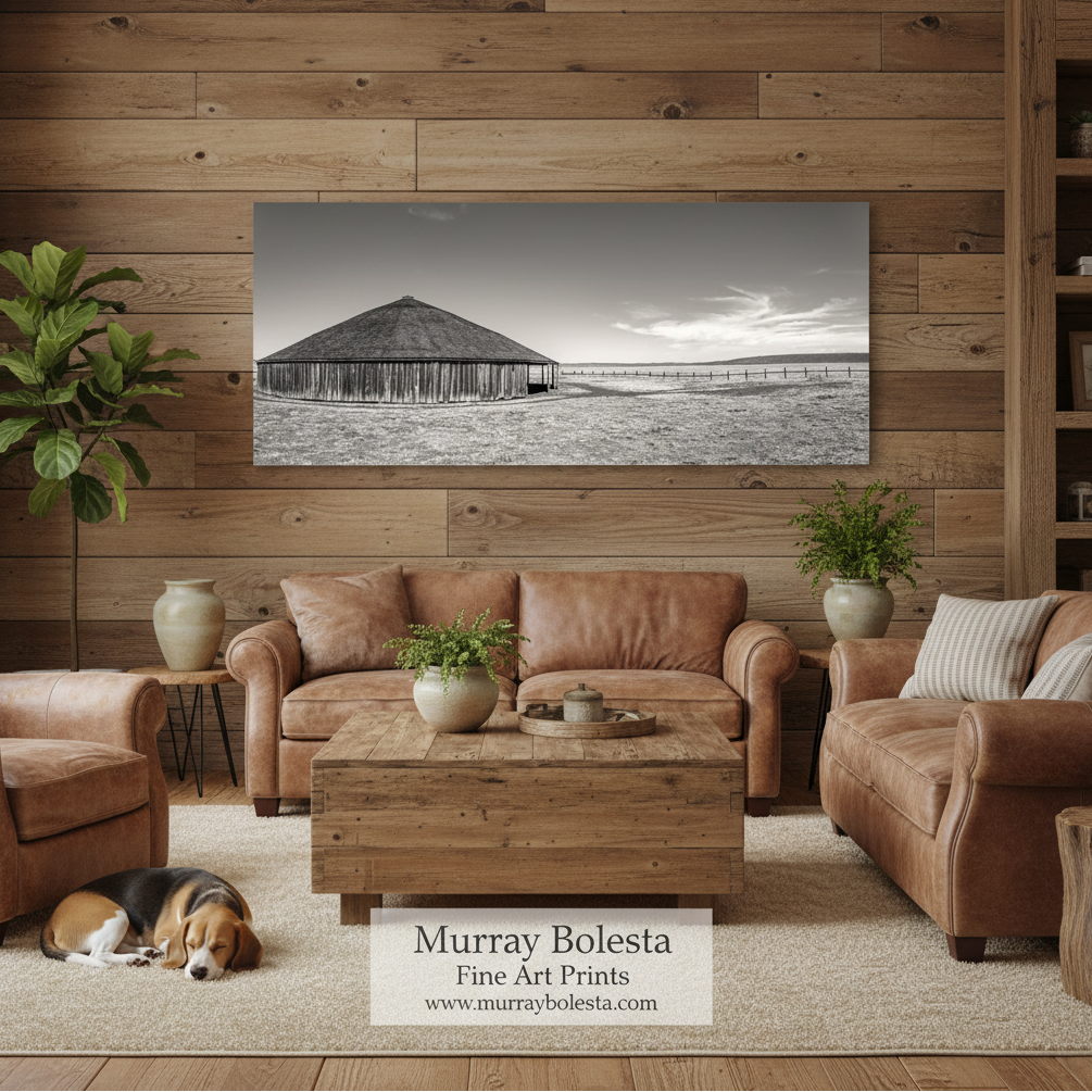 Oregon round barn! A sepia landscape art print of a wooden barn is displayed on a rustic wood-paneled wall in a cozy living room setting with brown leather sofas, a wooden coffee table, and a dog sleeping on the rug. A watermark at the bottom reads 'Murray Bolesta Fine Art Prints'.