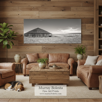 Oregon round barn! A sepia landscape art print of a wooden barn is displayed on a rustic wood-paneled wall in a cozy living room setting with brown leather sofas, a wooden coffee table, and a dog sleeping on the rug. A watermark at the bottom reads 'Murray Bolesta Fine Art Prints'.