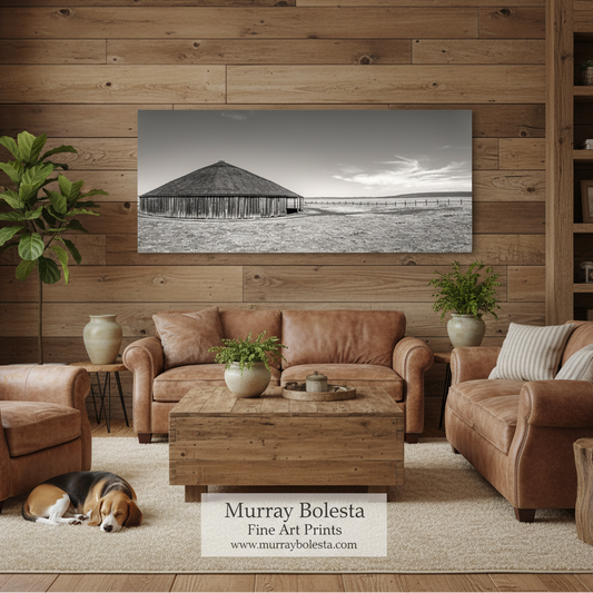 Oregon round barn! A sepia landscape art print of a wooden barn is displayed on a rustic wood-paneled wall in a cozy living room setting with brown leather sofas, a wooden coffee table, and a dog sleeping on the rug. A watermark at the bottom reads 'Murray Bolesta Fine Art Prints'.